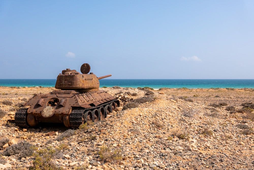 A rusting tank sits on the seashore, its gun pointing out toward the blue sea.