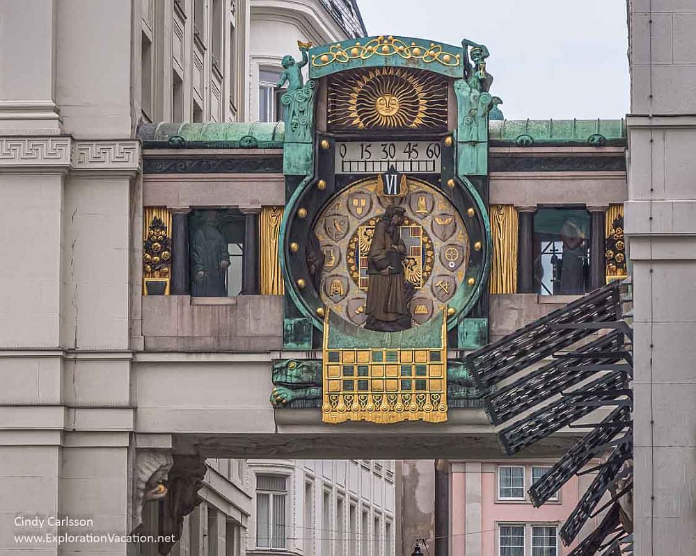 Jugendstil clock on a building's bridge over a street: golden images instead of numbers, a golden sun above.