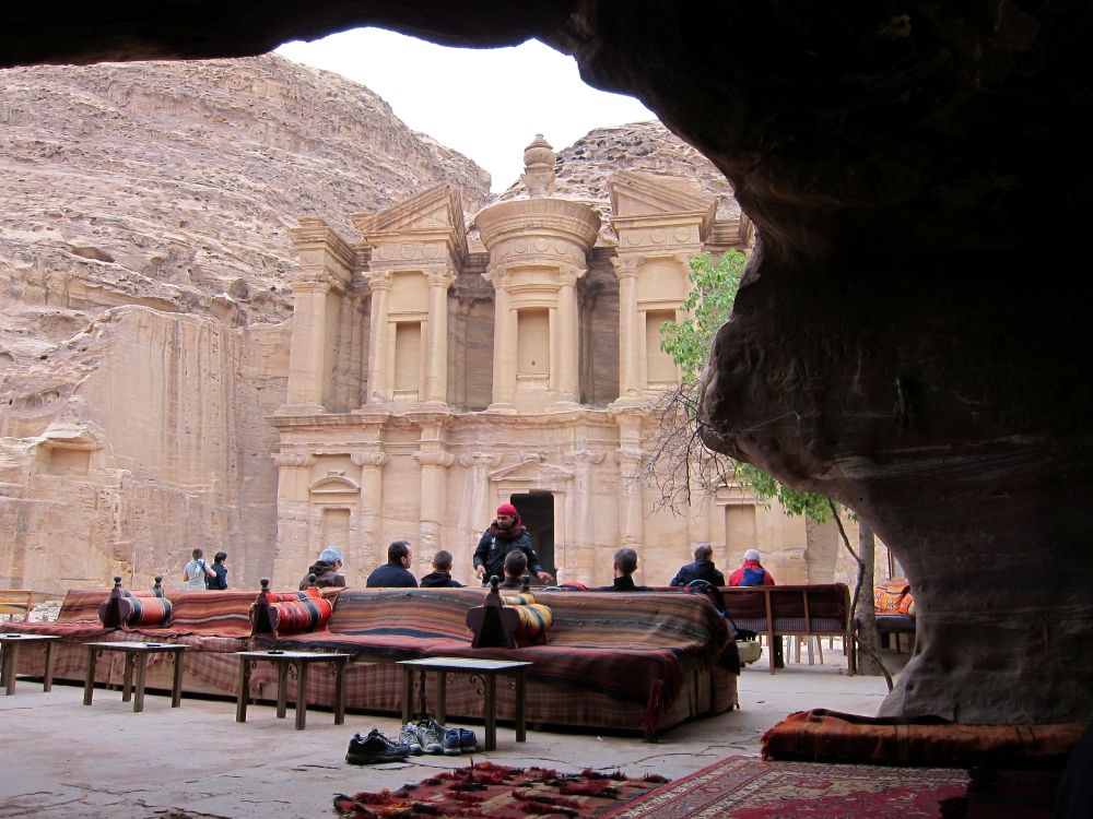 Seen from the opening to a cave, the monastery is carved from a cliff, with 3 towers (attached at the back still) and a single doorway in the center. In the open space in front of it, people sit on Bedouin-style sofas.