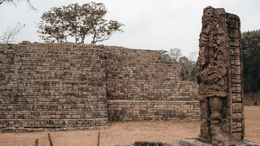 Ruin of a large stone building, perhaps a step pyramid, with in the foreground a stone statue of a human but with a very ornate headdress..