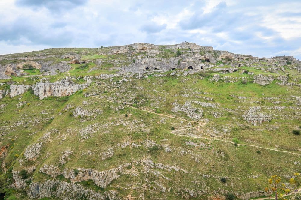 A grassy hill, with a path zigzagging up it, and cave dwellings near the top at the Park of Rupestrian Churches in Matera. 