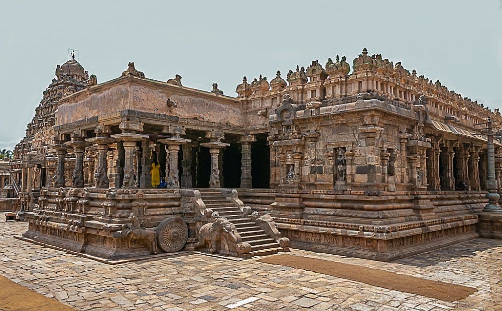 A large stone temple with carvings all around its roofline, steps up and a porch held up by carved pillars.