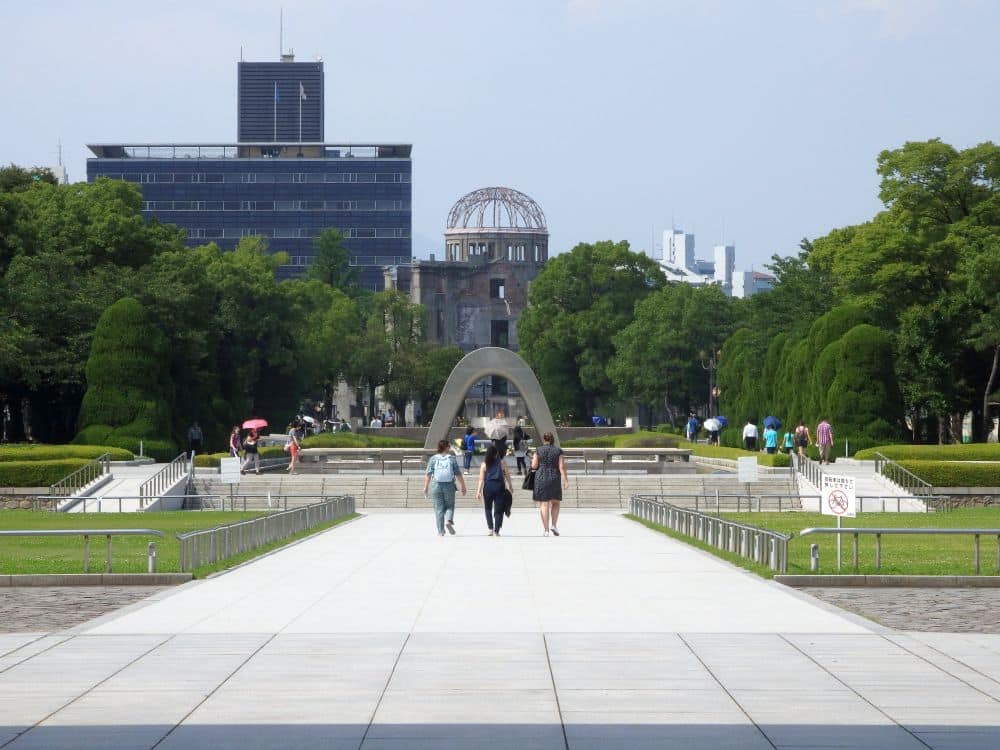 The arch of the cenotaph in the middle distance, with nearer to the camera, three people walking toward it. Directly behind the cenotaph is the Genbaku dome. On ether side, green lawns and trees.