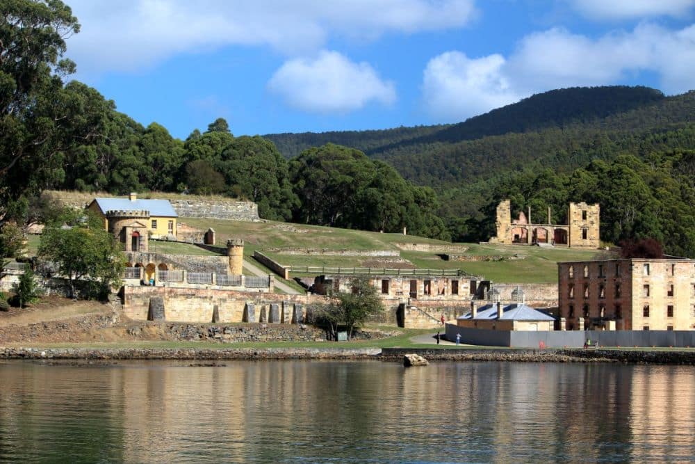 Seen from the water, several houses on a gentle slope, some intact, some ruins, at Port Arthur Historic Site, one of the Australian Convict Sites.