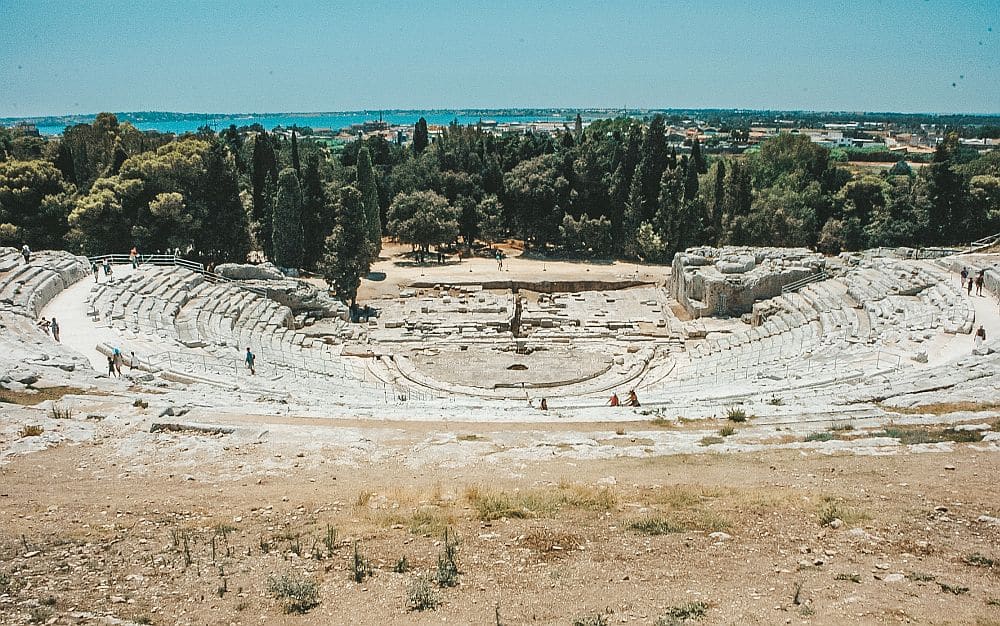 Looking down a slope at a large amphitheater with a semi-circular ring of rows of stone seats around a semi-circular stage at the bottom center.