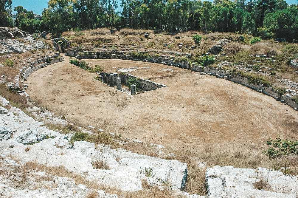 Limited remains of an oval amphitheater, with only the outlines of the original seating on the sides, and a big rectangular hole in the center floor.