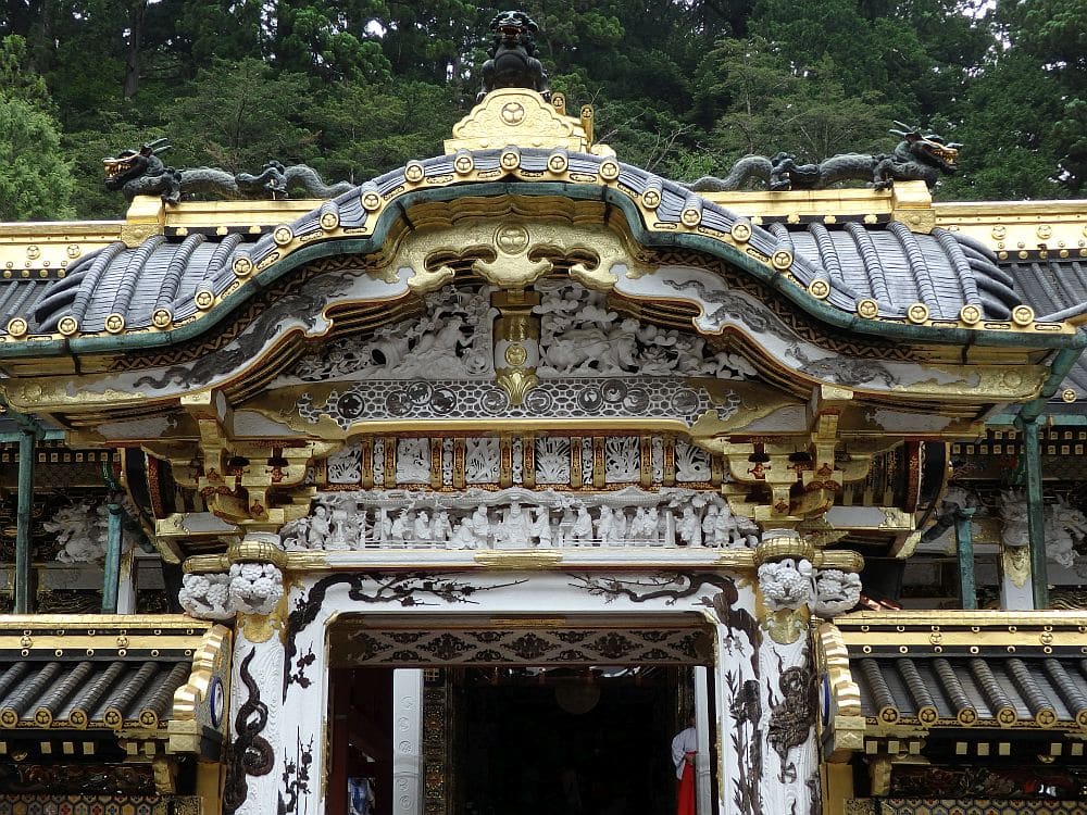 The photo shows only the area above the door of the gate of the shrine: under the eaves are ornate little bas reliefs depicting people and nature: most are white, but the roof elements around them are painted gold.