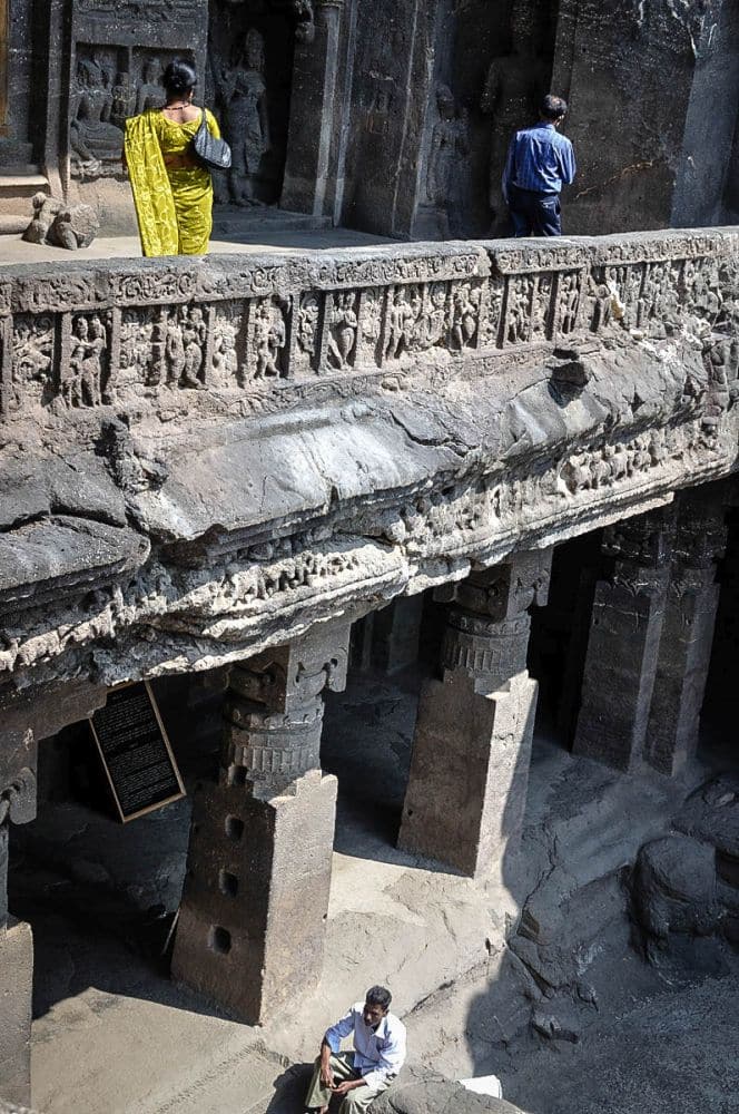 Taken from the second story of one of the Ellora Caves, there is a carved balustrade along a balcony, which is held up by carved heavy pillars. A woman in a sari stands on the balcony, back to the camera.