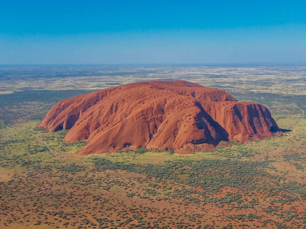 Aerial view: the massive red rock stands out from a perfectly flat, scrubby green and brown landscape.