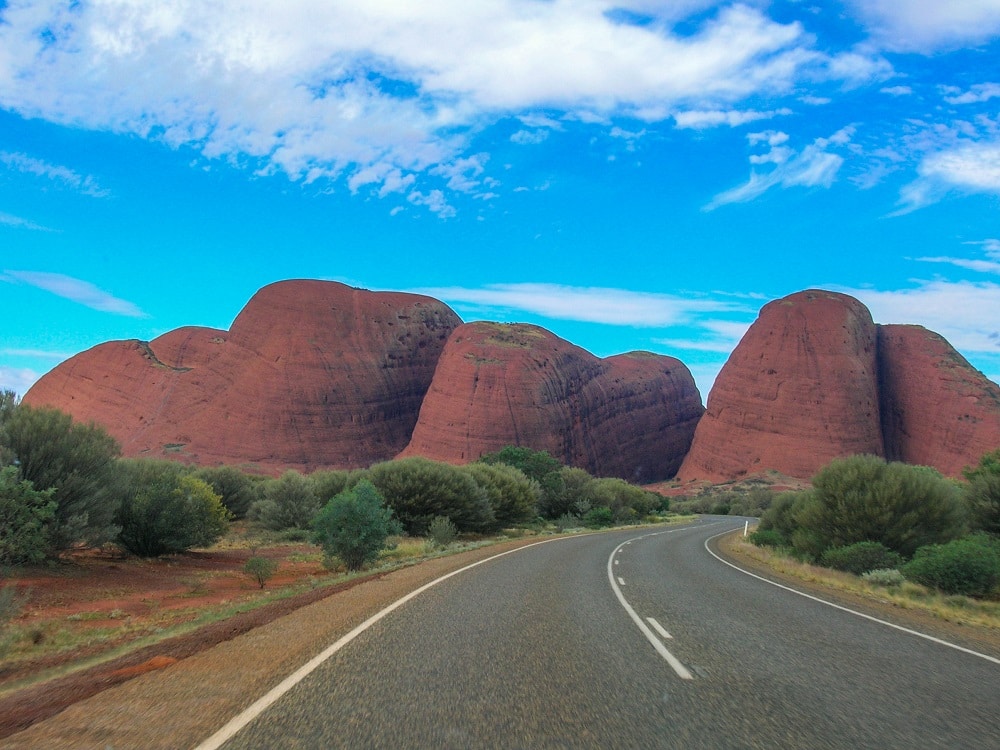 View down a roadway, with the rounded red-rock hills of Kata Tjuta in the distance.