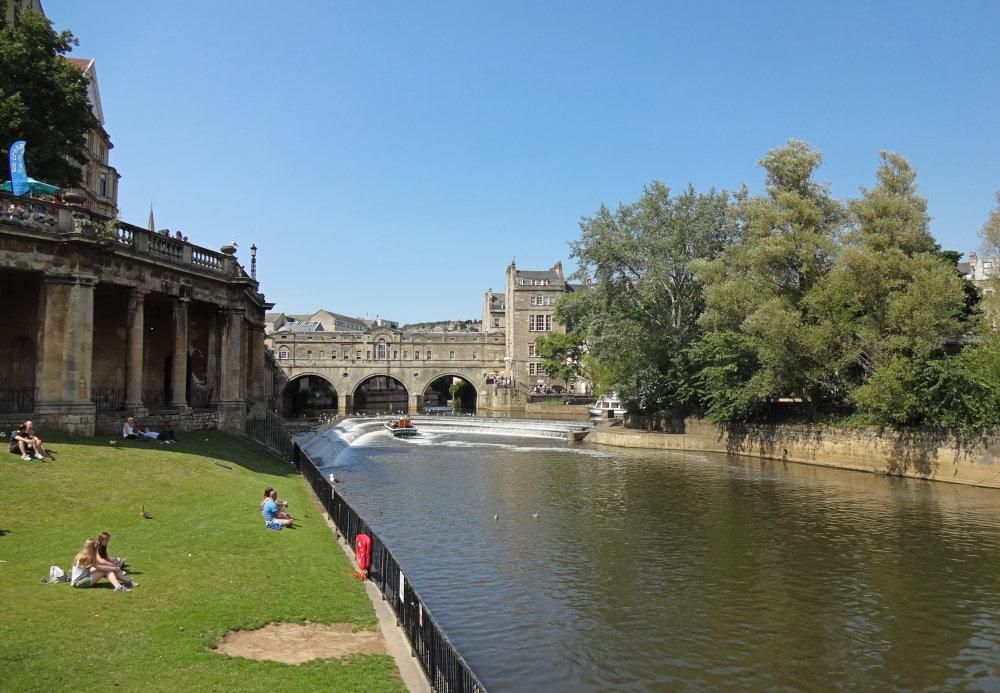 Looking down a river, with grass on the near bank, a bridge visible in the distance with arches over the water and buildings integrated into the bridge.