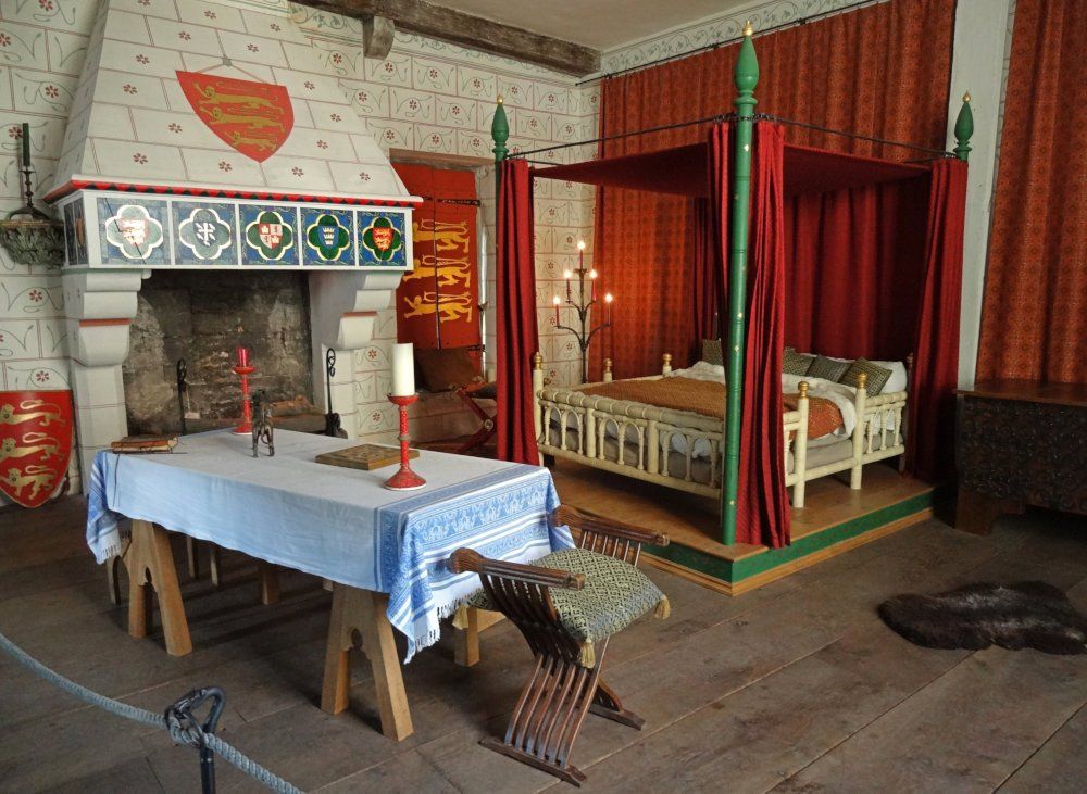 Interior view of a bedroom with a bed with a canopy, a long table and a large, decorated fireplace.