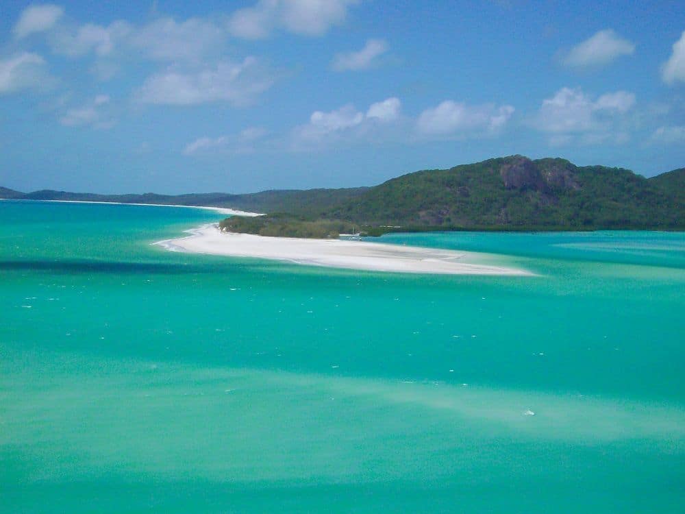 View toward land from the water: greenish-blue sea, and a point of land that ends in a flat white sand bar.