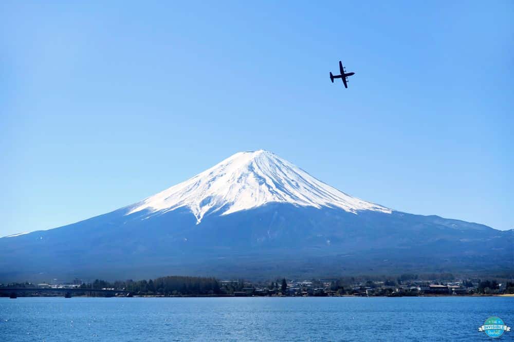 A view of snow-capped Mount Fuji across Lake Kawaguchiko, with a small plane above the lake.