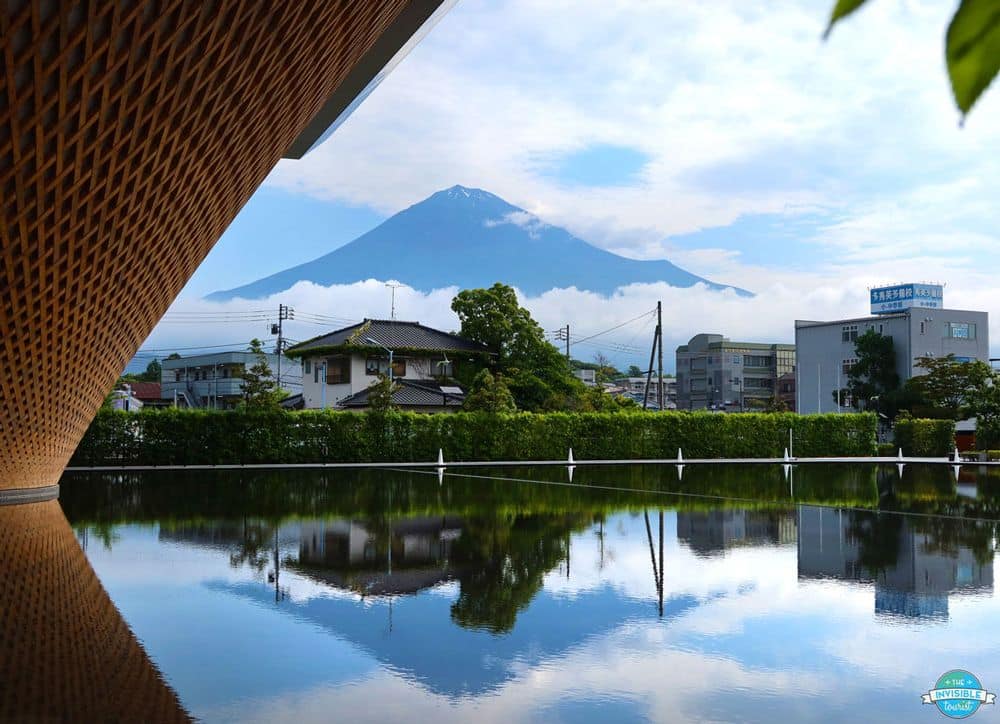View of Mt Fuji from the Mt. Fuji World Heritage Center in Fujinomiya: a lake, some buildings across the lake, and behind them, the cone of the mountain, with cloud around its base.