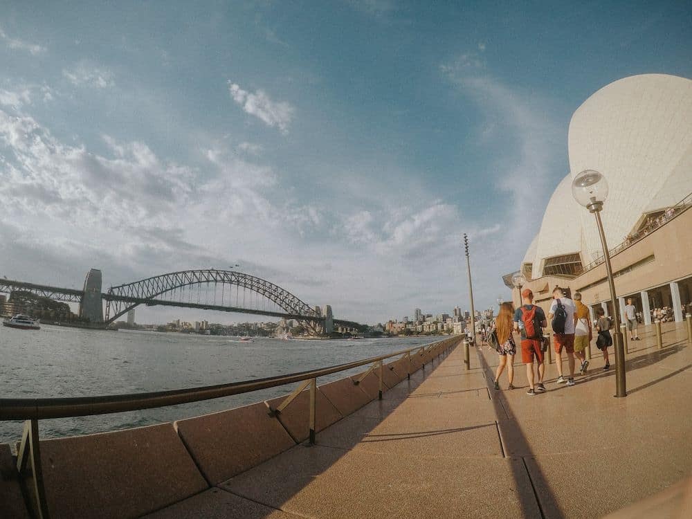 The side of Sydney Opera House on the right, a walkway straight ahead, and the water on the left, with a bridge in the distance.