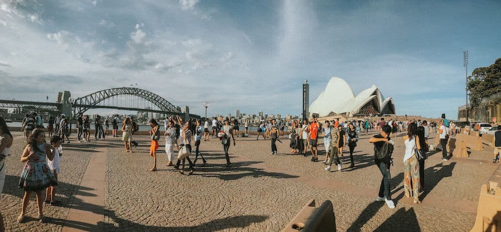 In the background, the Sydney Opera House on the right and a bridge on the left. In the foreground, lots of people strolling.