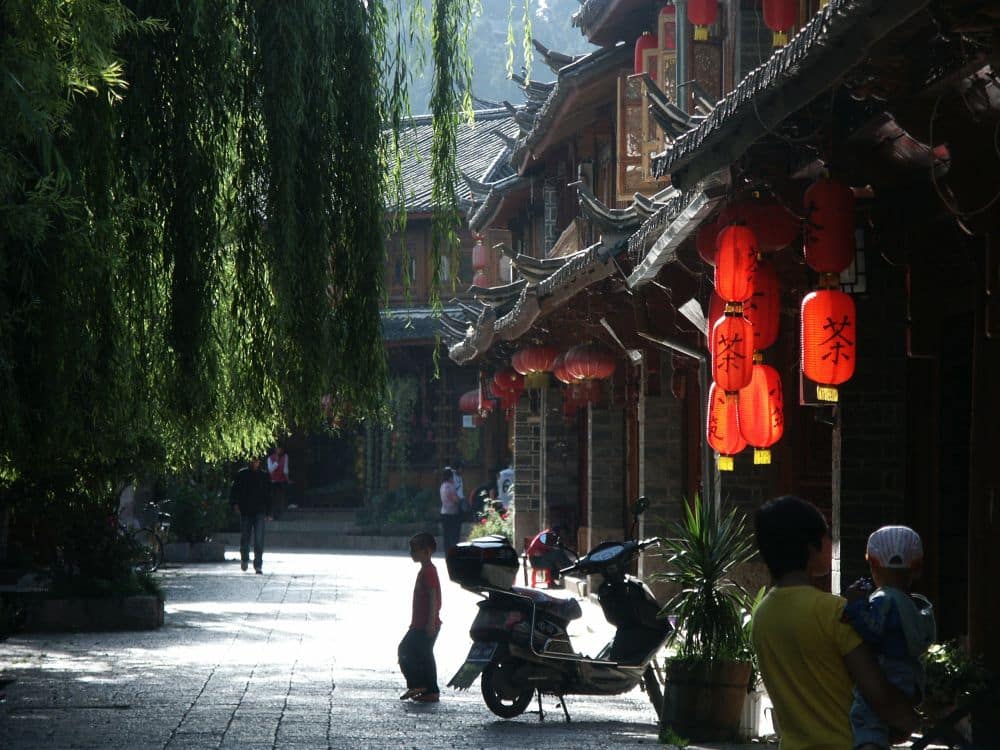A street in Lijiang: shop houses along the right, with red lanterns hanging from the eaves. Motorbikes parked, a child walks in the street, willow trees overhang the street on the left.