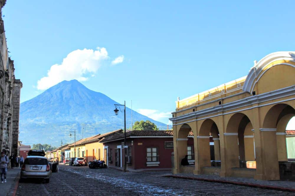 A view down a city street, with single-story shops along the sides and the cone of the volcano looming in the distance.