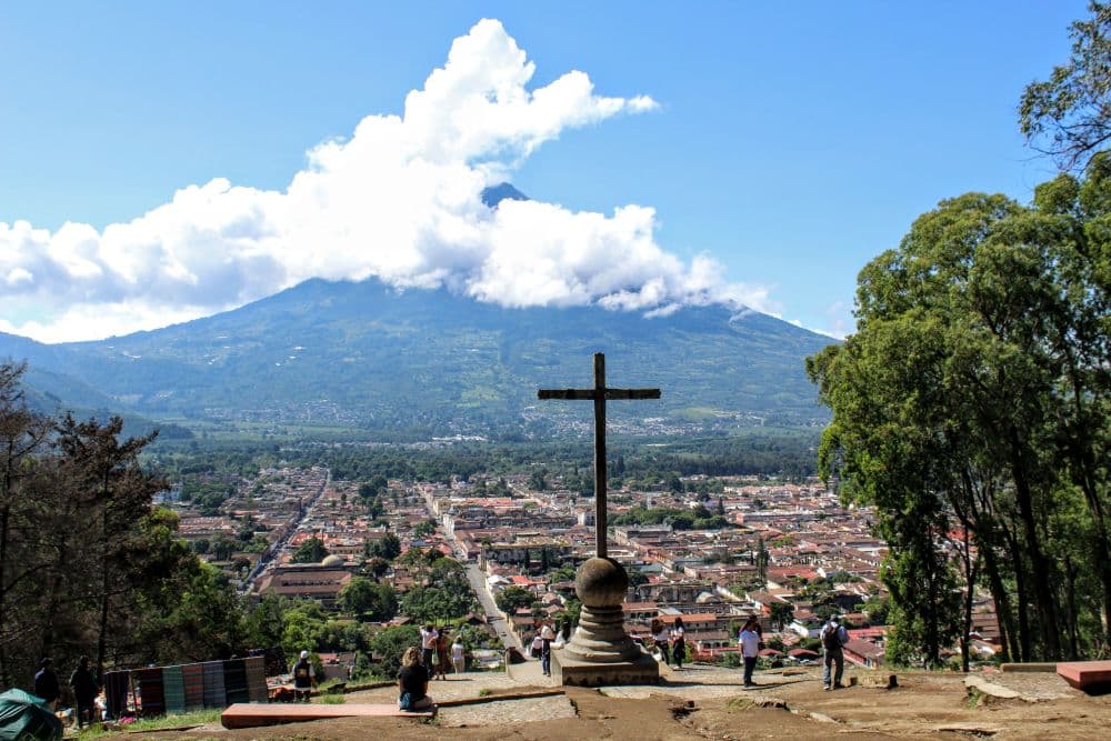 View over the city. In the foreground, a large monument with a cross. Below, the city, with its grid of streets, and in the background, the volcano, its peak surrounded by clouds.