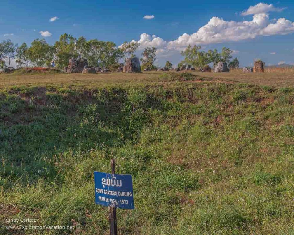 On the upper part of the photo, a flat grassy place with a scattering of stone jars. In the foreground, a steep grass-covered section with a sign reading "Bomb craters during war in 1964-1973"