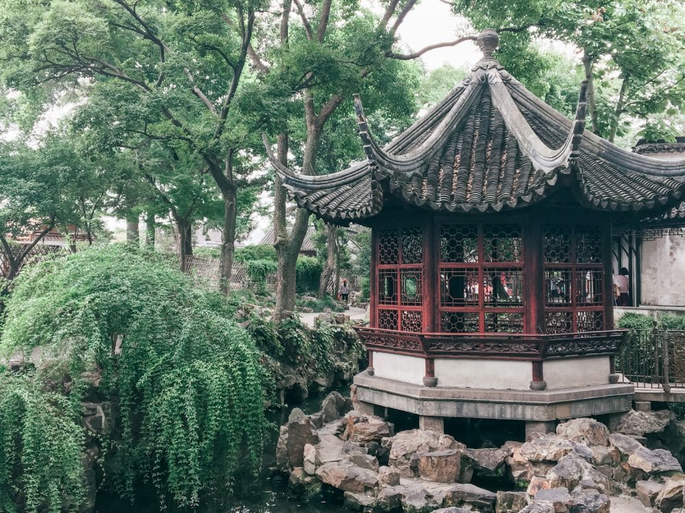 A small round pavilion with a typical curved Chinese roof stands on stones above a pond with various trees behind it.
