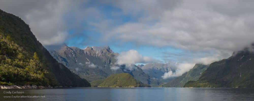 A body of water with, around it, very picturesque mountains, clouds hovering above.