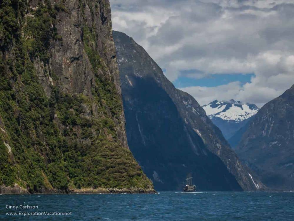 Mountains rise steep from the water, and a 3-masted ship looks tiny next to them.