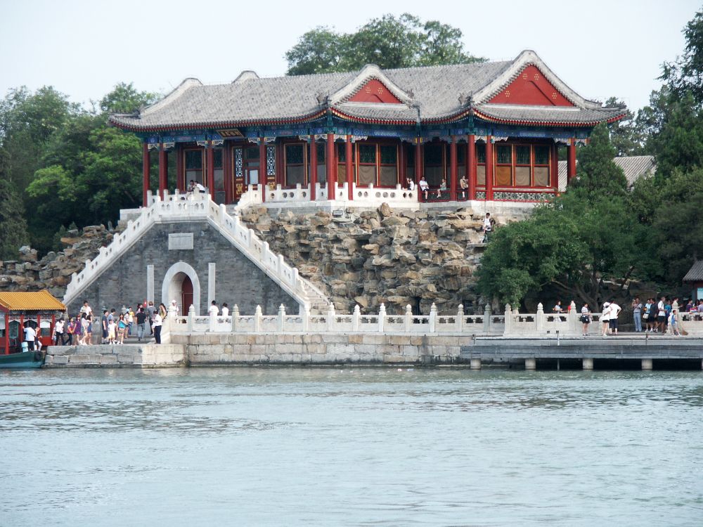 Seen across the water, a walkway long the edge of the water and, above it on a higher level of land, a red temple with curved eaves.