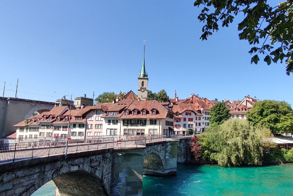 View of a bridge across a river leading to a pretty town of white buildings with red roofs, a steeple peeking up above them.