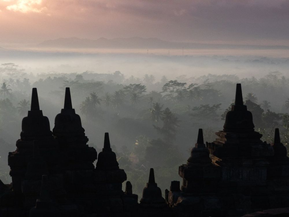 After sunrise, the pointed stupas of Borobudur silhouetted against the forest below as the sun rises.