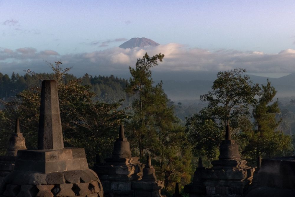 A view from the temple: stupas in the foreground, forest in the middle ground, and a volcanic mountain peeking pu above a layer of cloud in the background.