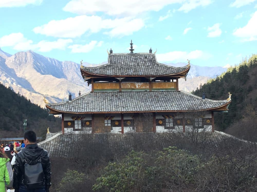 A small temple, brown and white wood with grey roofs with curved roof lines. Mountains in the distance.