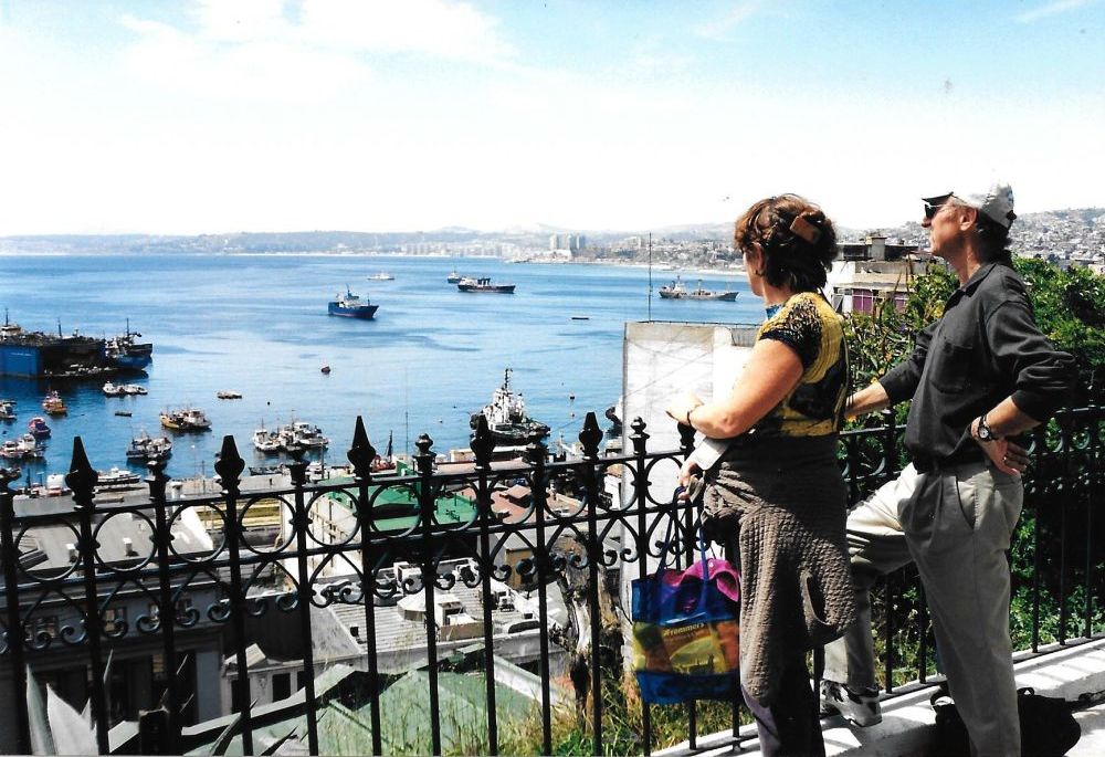 Image: two people stand at a fence overlooking a view of the city and the sea.