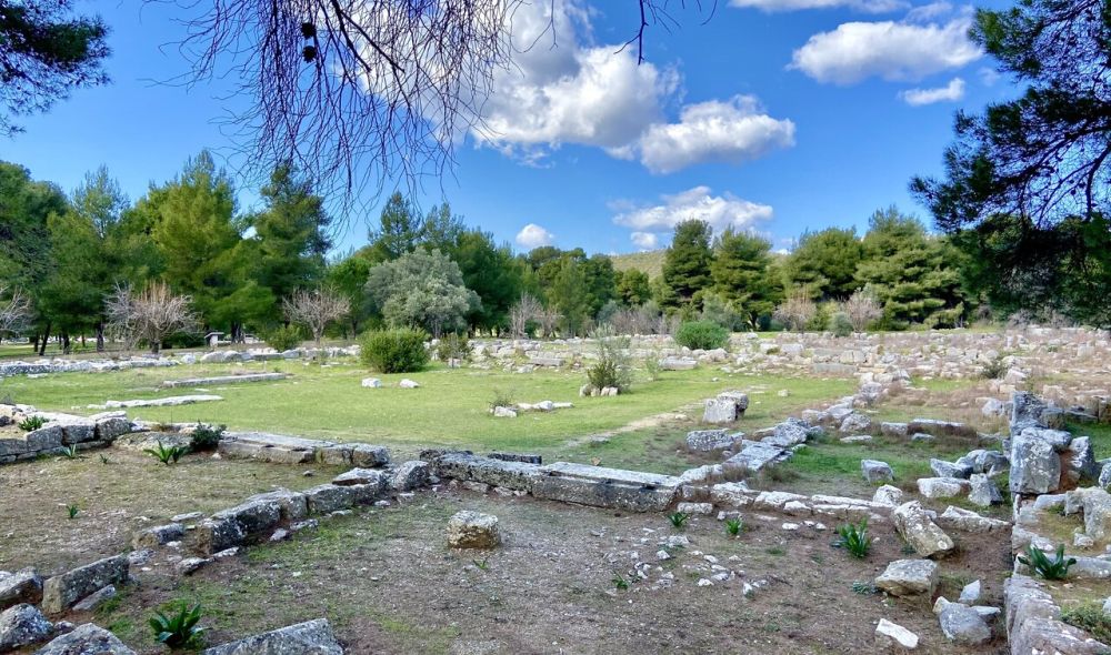 A flat field with trees around its edge and low stone walls marking out where the original rooms were.