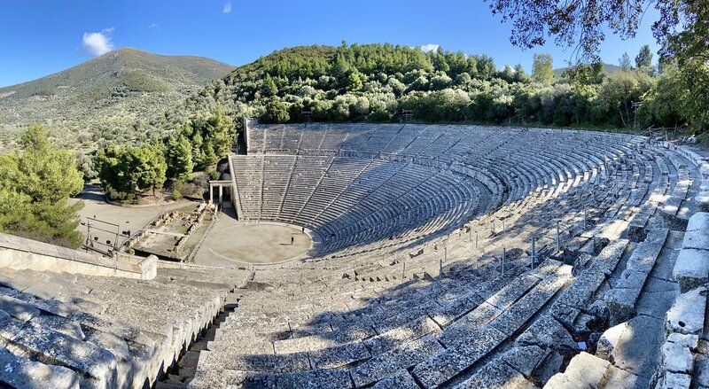 The theater, seen from one side, is concentric half-circles of stone seating around a semi-circular stage at the bottom.