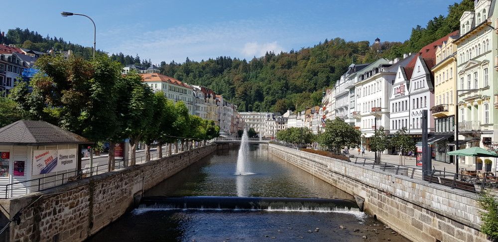 View down a tree-lined canal with a fountain in its middle and rows of neat buildings on each side.