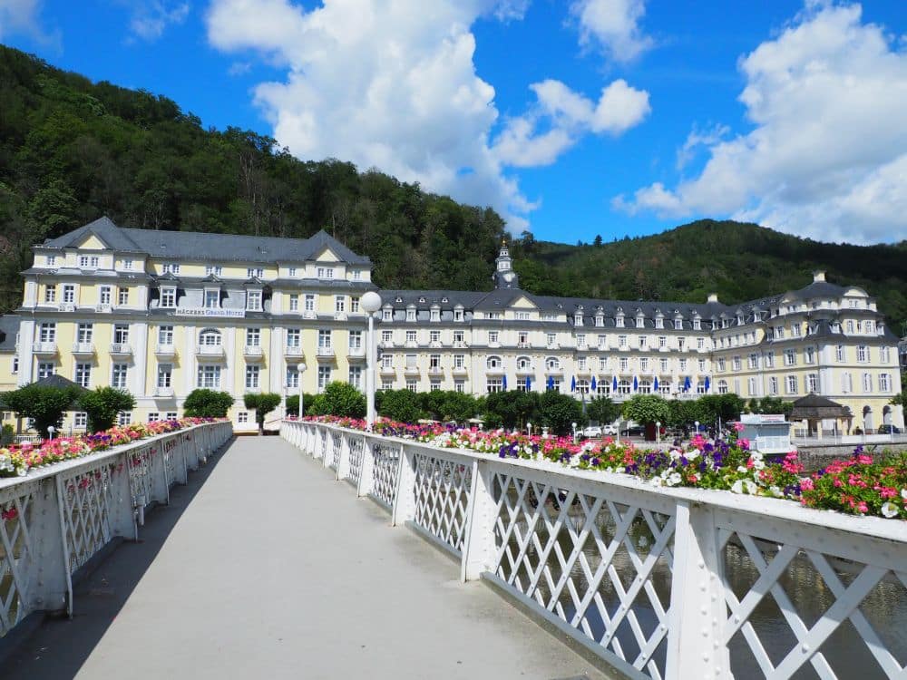 View along a bridge, white rails and flower boxes on each side, leading to a large, palatial building in pale yellow.