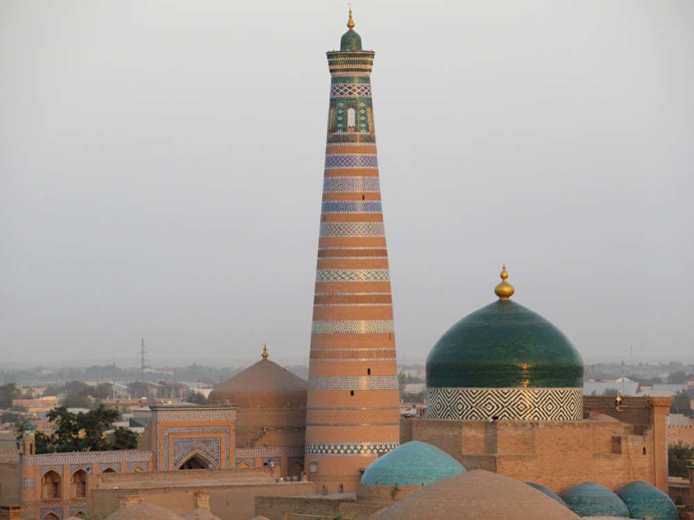 The minaret is very tall with horizontal decorative bands around its circumference. It is much wider at the bottom than the top. A large green dome covers the mosque next to it.