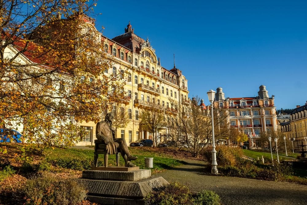 Grand-looking neo-classical buildings surround a park, with a statue in the park of a man sitting in a chair.