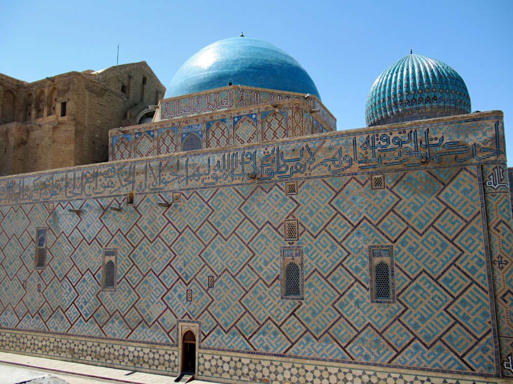 View of the side of the structure: flat wall with decorative tiles, a small door at ground level and a few small windows. Above, two tile-covered domes.