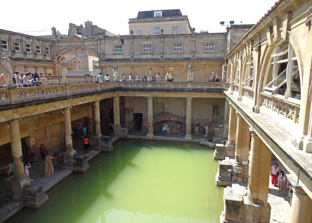 Looking down from a balcony above the green water of the Roman Bath. The lower floor around the pool has a colonnade around it, held up by pillars.