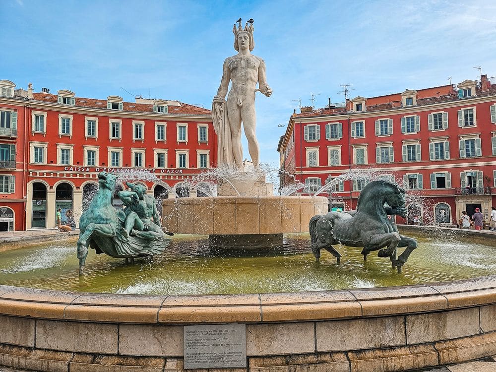 A fountain in the center of Place Massena: statue in the center, pretty red buildings behind.