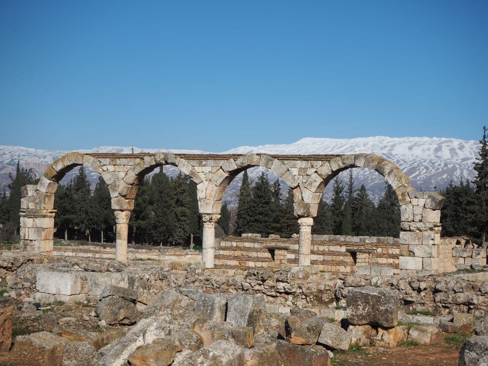 View across part of the site to a row of 5 pillars holding up arches. Beyond the arches, snow-covered mountains.