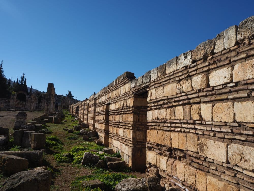 Looking along a row of walls about 6 ft or 2 meters high with doorways at regular intervals. The wall is made of alternating horizontal rows of stone and bricks.