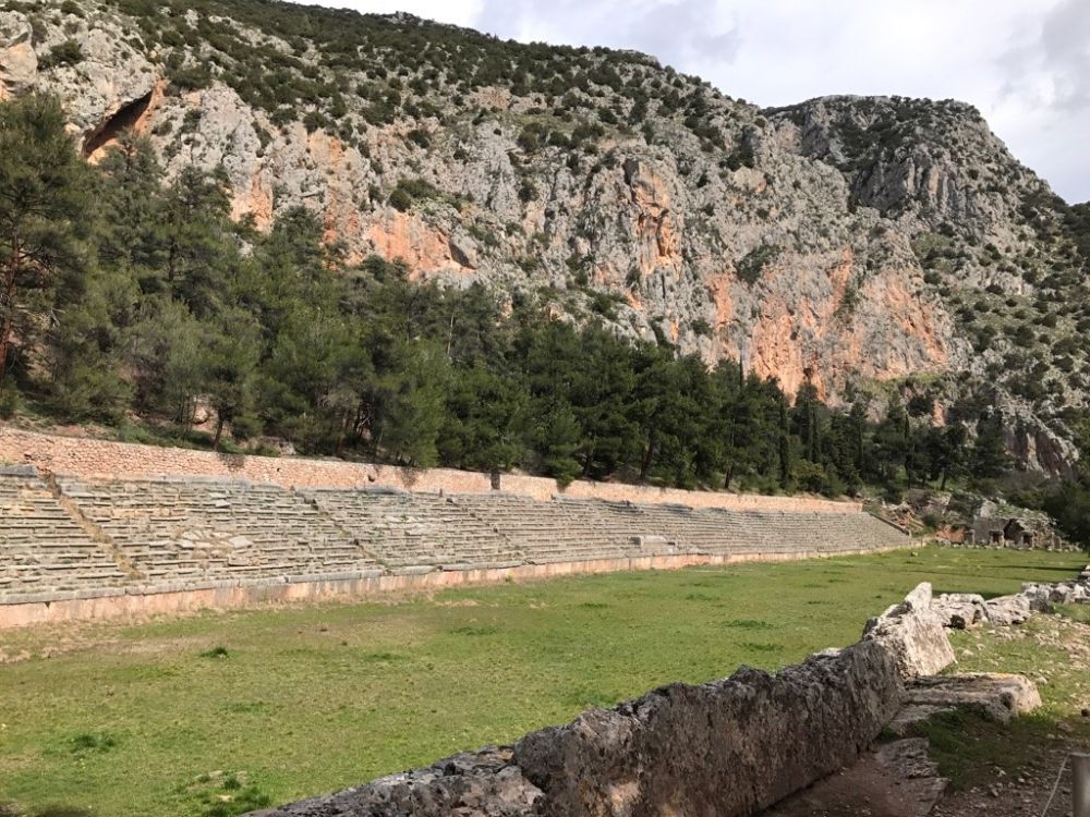 Looking down the long narrow stadium: bleachers of stone on the sides, a grassy center floor. Behind rises a mountain.