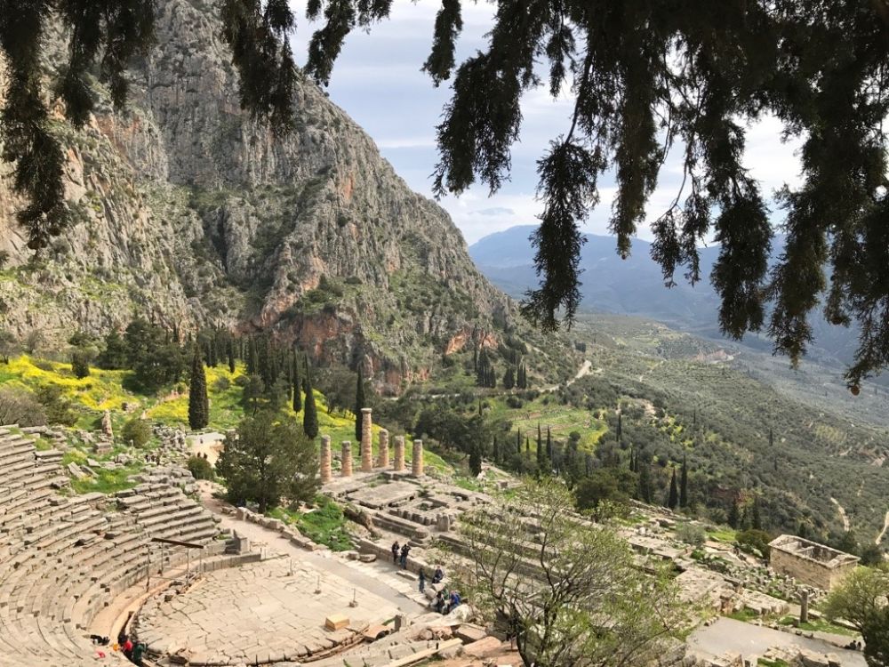 View down a hill with mountains in the background. On the hill below some of the ruins are visible: the amphitheater and some columns and low walls.