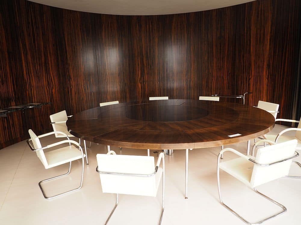 The table is dark brown and round, with 8 white chairs around it. The wall behind it is dark brown and curved in line with the curve of the table and richly grained.