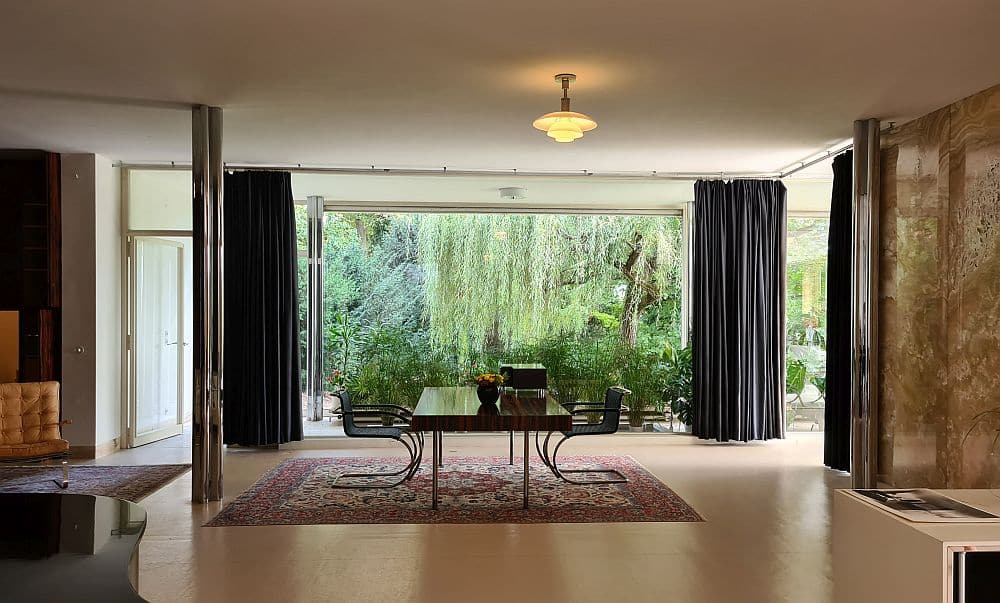 A view of a large room, a table and chairs at the other end of the room and a large glass wall behind, where a row of plants is visible in front of the outdoor trees.