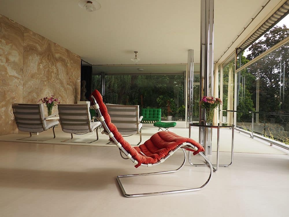 Looking from the dining area into the living room area: a chic red lounge chair in the foreground, leather and chrome; other chairs beyond that, with the onyx wall on the left and full height glass walls straight ahead and to the right.